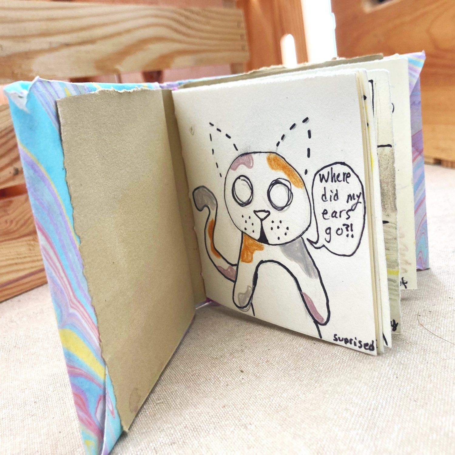 An illustrated book with a marbled cover sits open on a beige surface, revealing a charming drawing of a cat looking puzzled and asking, “Where did my ears go?!” Perfect for cat lovers and fans of whimsical art, this unique book draws readers in with its playful illustrations and beautifully designed cover.