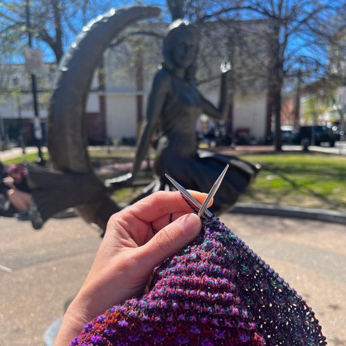 In the foreground, a vibrant hand knitting with purple yarn steals the spotlight, while in the background, a captivating bronze statue of a woman poised on a crescent moon adds intrigue. Framed by lush trees and charming buildings under a bright blue sky, this scene beautifully blends creativity and culture.