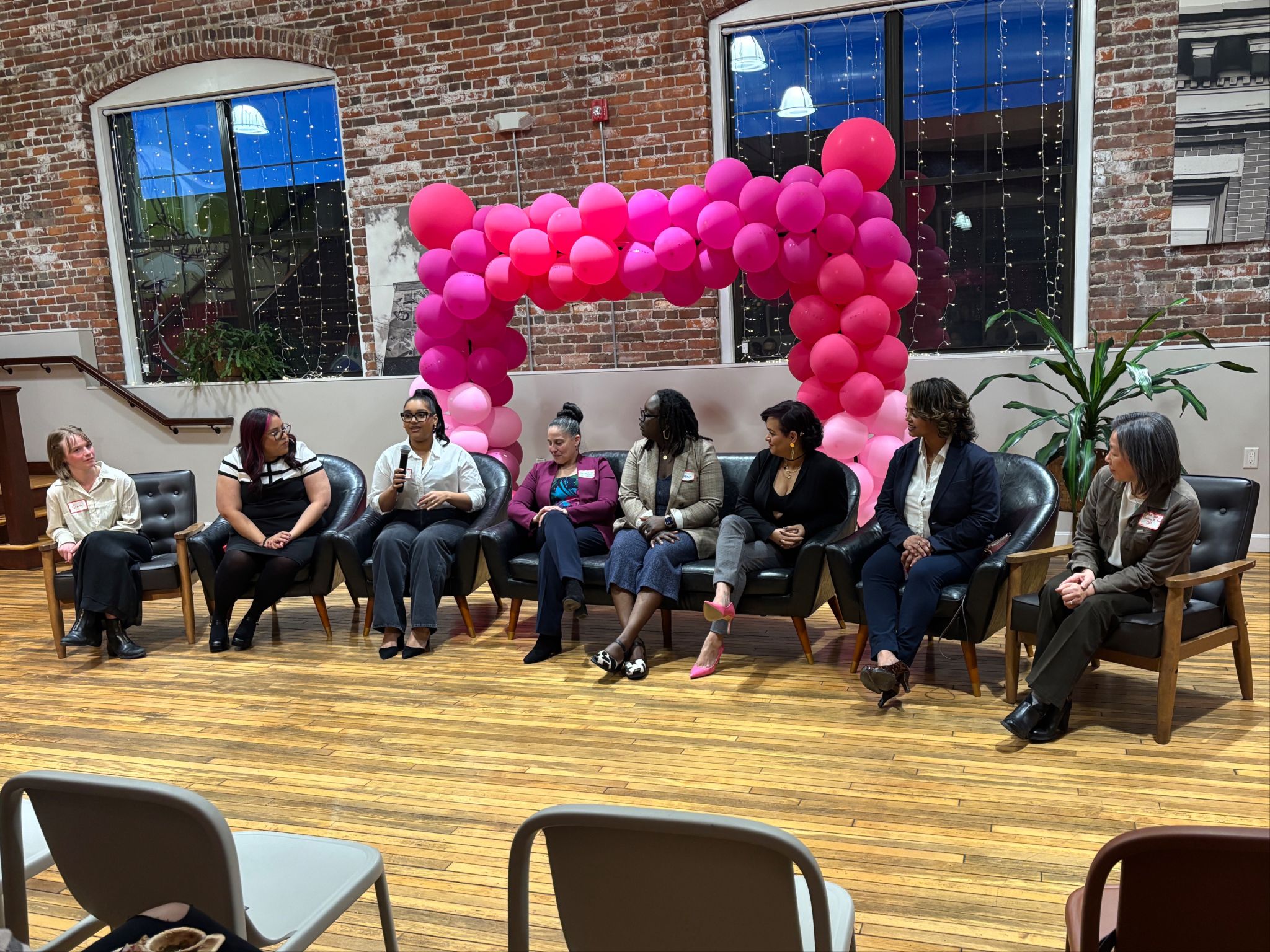 Seven women participate in a lively panel discussion, seated before a pink balloon arch. The event takes place in a stylish space with brick walls, wooden floors, and large windows, creating a warm and inviting atmosphere. Audience members can be seen listening attentively to the conversation.