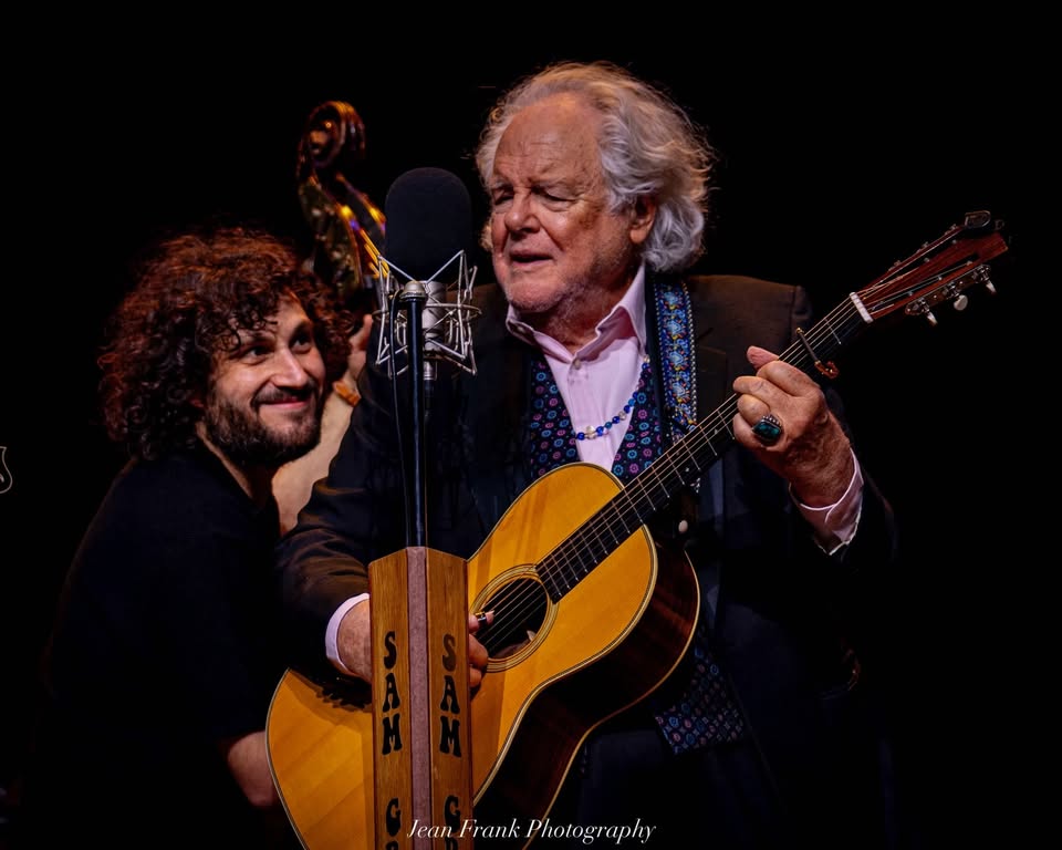 An older gentleman in a suit plays guitar and sings on stage, joined by a younger man with curly hair who smiles while holding a double bass. Together, they deliver a lively live music performance that captures the crowd’s attention.