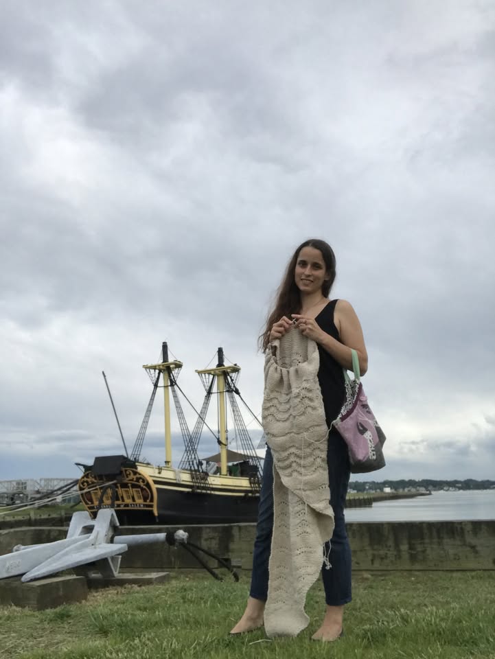 A barefoot woman stands on the grass near a dock, wrapped in a cozy knitted blanket. Behind her, a striking historic tall ship with yellow and black masts creates a dramatic backdrop against the cloudy gray sky.