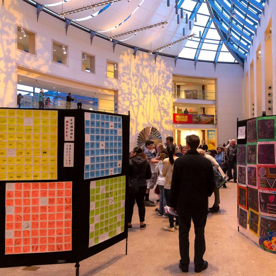 Visitors explore vibrant art displays inside a spacious, sunlit atrium. Natural light streams through the glass ceiling, casting striking tree-like shadows on the walls. Bright spotlights highlight each artistic piece as crowds gather to admire the colorful exhibits in this modern setting.