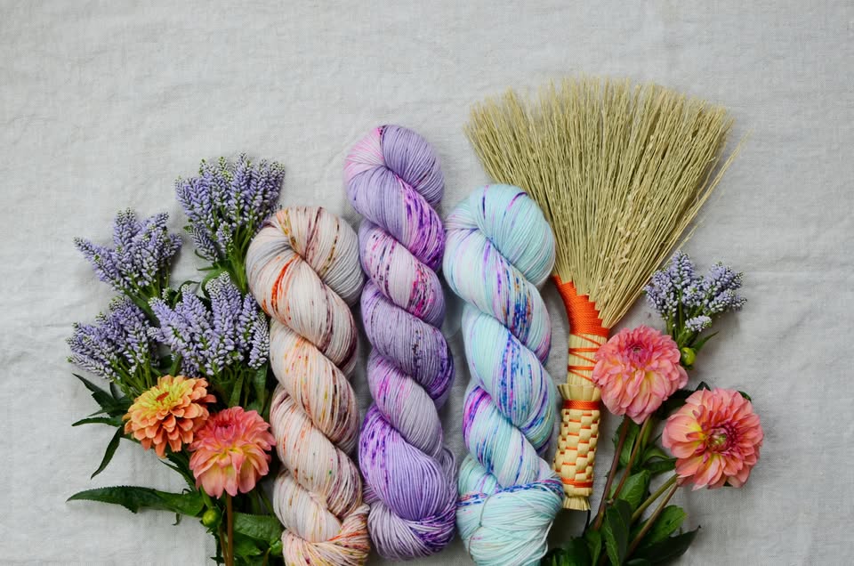 Three vibrant skeins of multicolored yarn sit neatly in a row, framed by purple and pink flowers, with a charming straw broom accent, all set against a soft fabric backdrop. This eye-catching setup brings color and texture together—perfect for craft lovers and those seeking creative project inspiration.