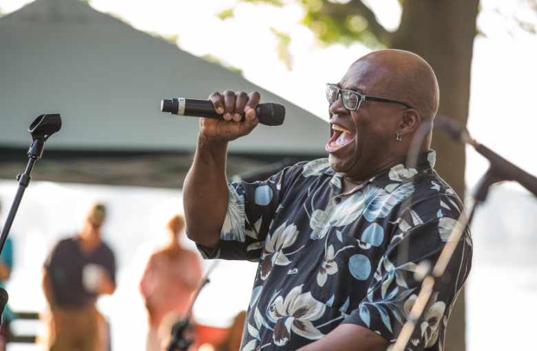 A man in glasses and a floral shirt sings energetically into a microphone at an outdoor event, capturing the attention of a crowd beneath a canopy. The lively scene is filled with blurred guests in the background, highlighting the vibrant atmosphere of this open-air performance.