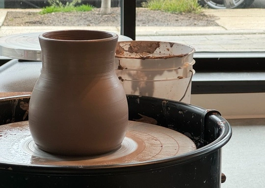 A newly crafted clay vase rests on a potter’s wheel, ready for finishing touches. In the background, a white bucket of slip and essential pottery tools sit near a window that overlooks the sidewalk and lush greenery outside. This peaceful scene captures the artistry of handmade ceramics in natural light, inviting creativity and inspiration.