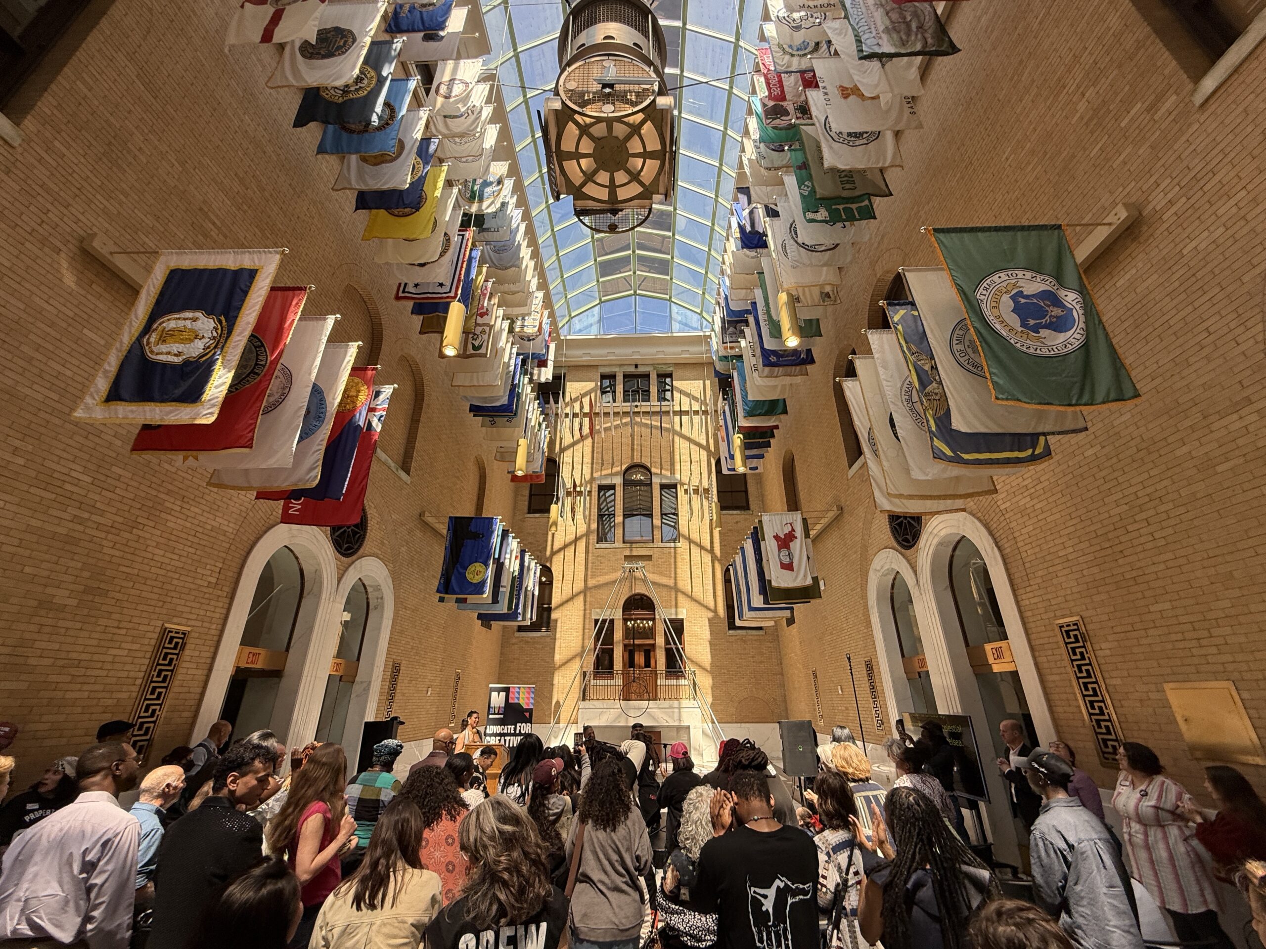 A lively crowd fills a bright, sunlit hall with a striking glass ceiling overhead. Sunny yellow brick walls surround the space, while rows of vibrant flags hang above, adding splashes of color. At the center, a large clock catches the eye as people turn their attention toward an inviting stage at the front—creating an energetic and welcoming atmosphere perfect for any event or gathering.