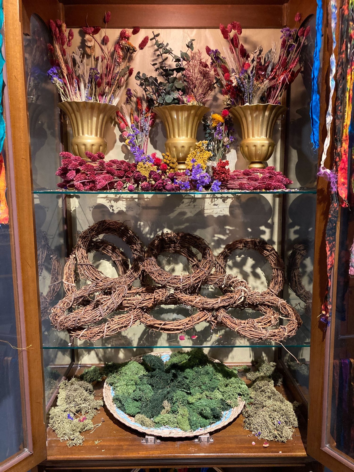 Discover the charm of a wooden display cabinet styled for instant appeal. The top shelf features elegant gold vases filled with dried flowers, adding a touch of warmth and color to your space. On the middle shelf, rustic grapevine wreaths create an inviting, natural feel. The bottom shelf is anchored by a large bowl heaped with lush green moss—perfect for bringing the outdoors in and making your home decor stand out.