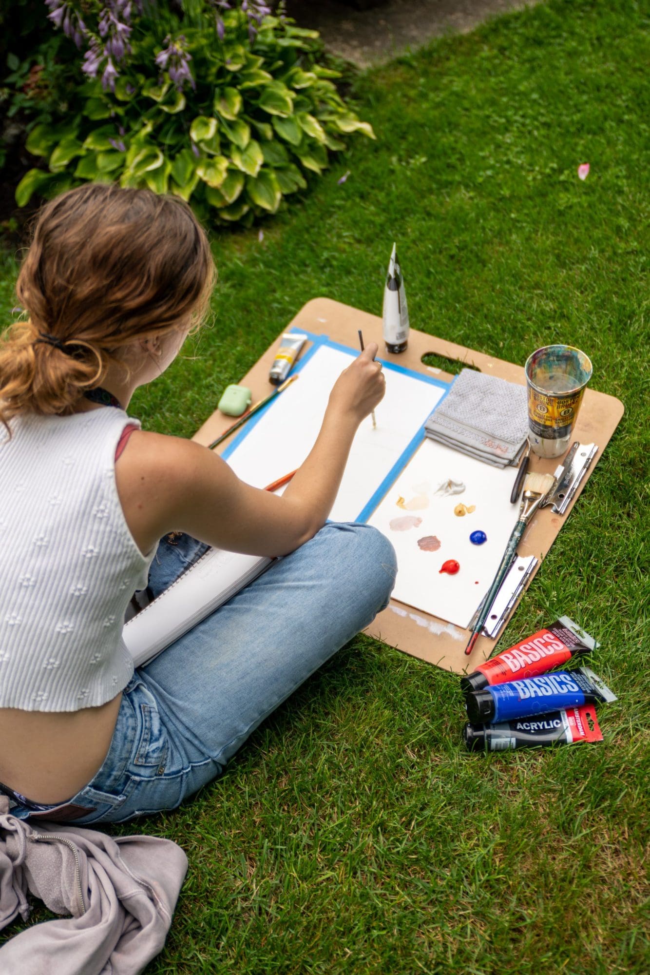 Sure! Here are a few revised alt text options based on your guidelines: 1. One of our artists paints outdoors on lush grass, surrounded by vibrant paint tubes, brushes, and blooming purple flowers. 2. We gather in the grass, sketchpads out and art supplies at hand, painting together among green plants and lively purple blooms. 3. Our creative community comes alive: someone painting on the lawn amid bright paint tubes and fresh local flowers. 4. A member of our creative crew paints in nature, sketchpad ready—art supplies scattered by colorful plants and purple blossoms. Each option describes what's visible while emphasizing creativity, energy, and community impact!.