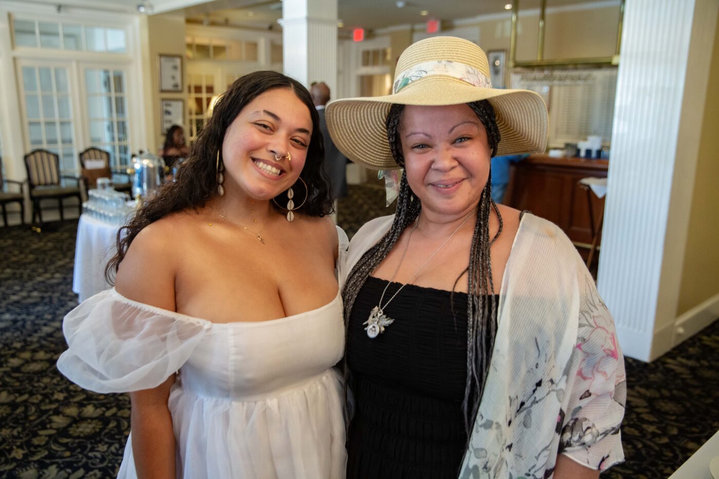 Two women indoors share a joyful moment, smiling and posing for the camera. One is dressed in a white off-the-shoulder dress with statement earrings, while the other pairs a black dress with a floral shawl and stylish wide-brim hat. Behind them, tables are set against an elegant backdrop, adding to the sophisticated atmosphere.