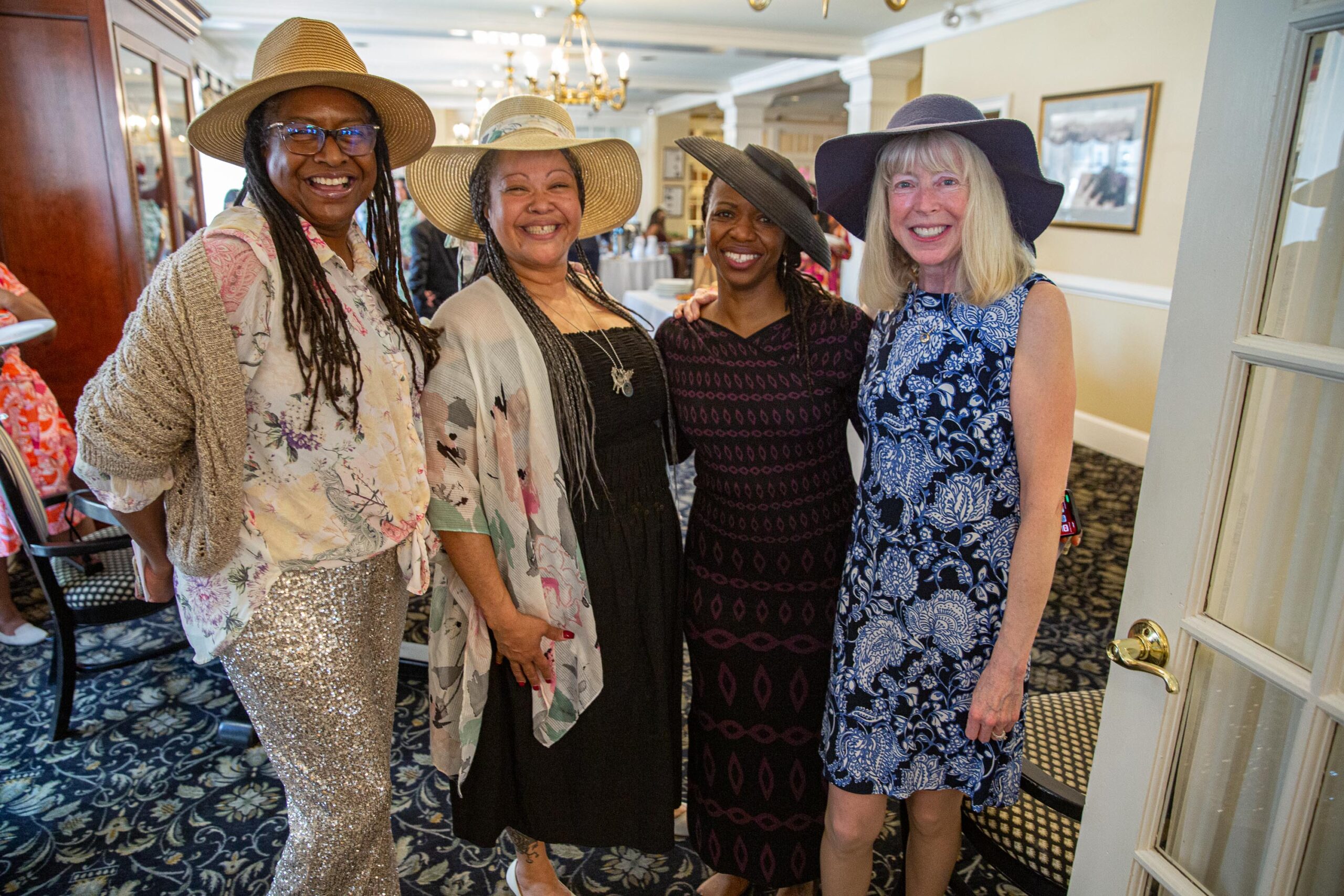Four women in fashionable hats and chic dresses share a joyful moment at an indoor social event. Standing close together and smiling, they radiate friendship and style against the backdrop of a bright, elegant room with a sparkling chandelier.