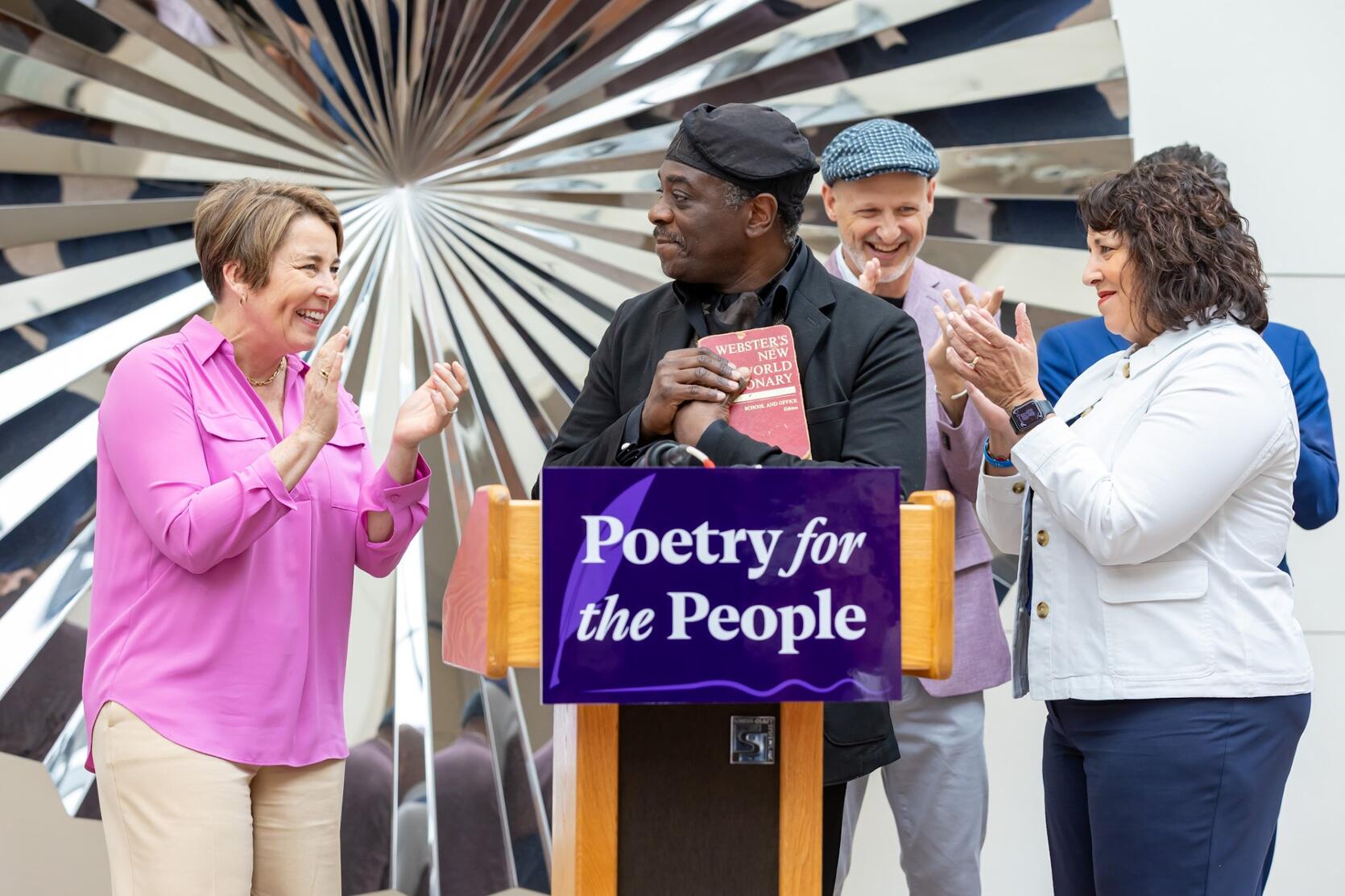 Four smiling adults gather at our "Poetry for the People" podium, cheering as we celebrate a local poet against a bright, artistic backdrop.