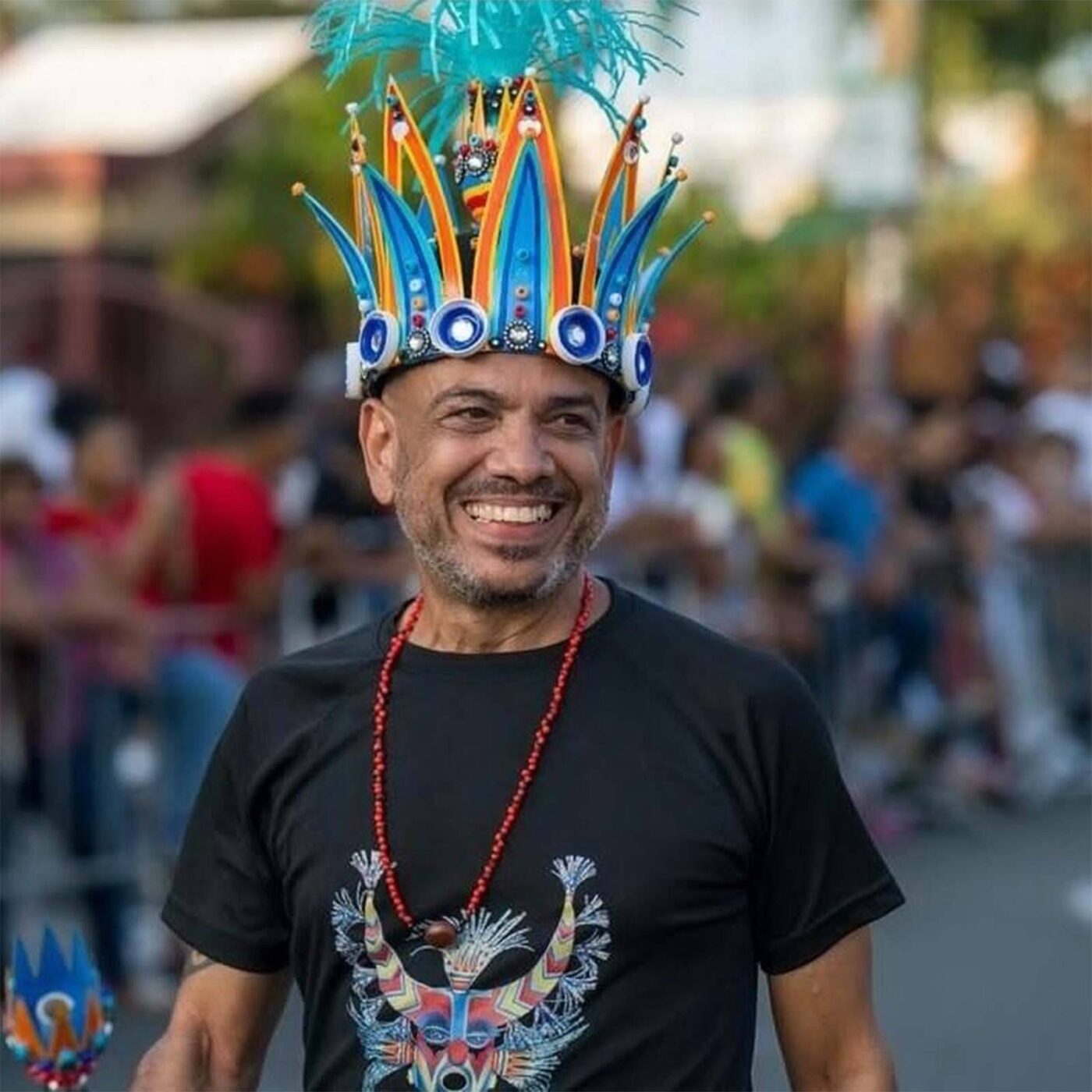 Smiling man in a bold blue and orange feathered crown, rocking our vibrant animal tee—celebrating local creativity with our community.