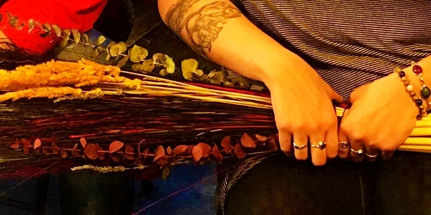A stylish individual wearing rings, beaded bracelets, and a striped shirt holds a rustic bouquet of dried flowers and leaves. Their forearm tattoo adds personality to the scene. In the background, someone dressed in red appears slightly out of focus, adding depth to the image.
