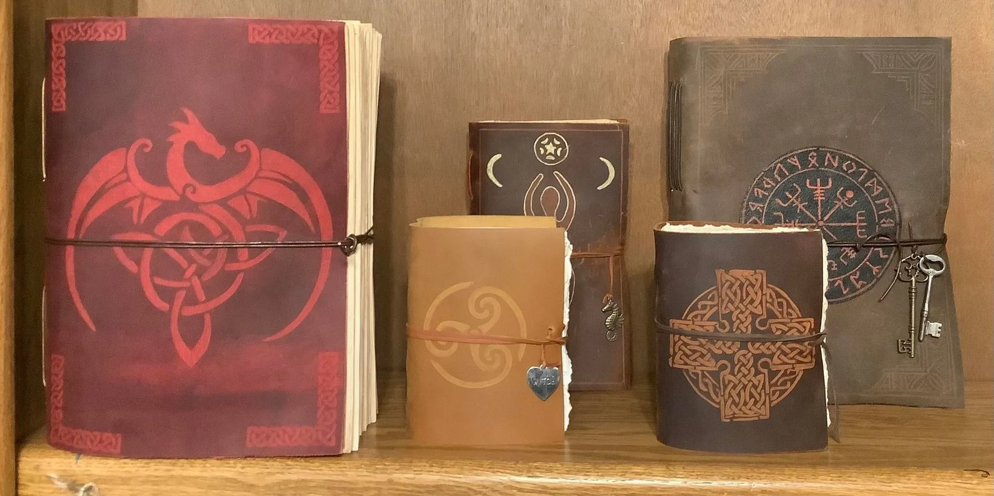 Discover five stunning leather-bound journals displayed on a rustic wooden shelf, each featuring intricate Celtic and Norse designs. One journal catches the eye in deep red with an embossed dragon, while the others come in earthy brown and tan shades, adorned with unique symbols. For a touch of mystery and adventure, a set of vintage keys hangs from the rightmost journal—perfect for anyone who values style and storytelling in their writing essentials.