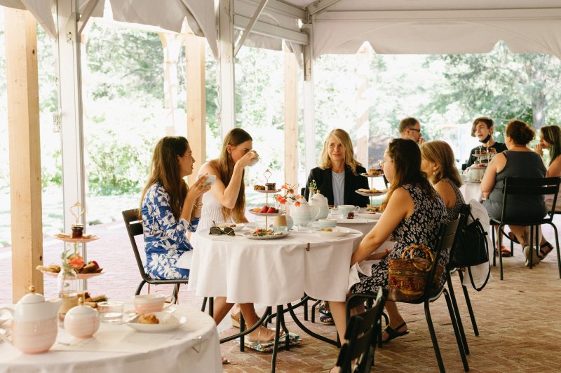 A joyful group of women in colorful summer outfits share tea and pastries under a white tent, surrounded by lively greenery—celebrating our vibrant community spirit.