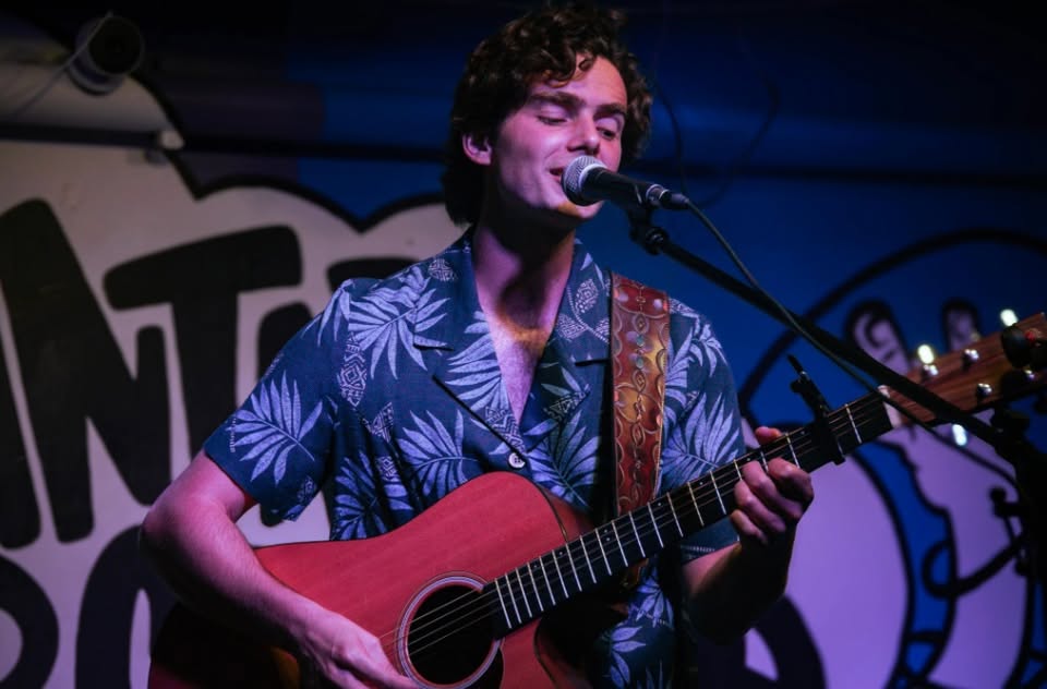 Sure! Here’s a more vivid, community-focused alt text under 125 characters: Our local musician rocks the stage—curly hair bouncing, blue shirt shining—surrounded by lively, colorful art.