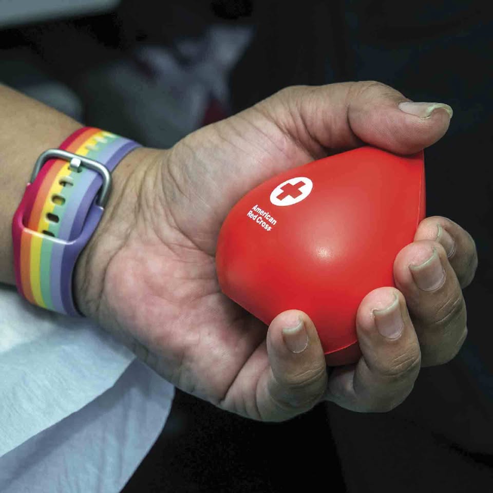 A hand in a colorful rainbow wristband squeezes a bold, heart-shaped Red Cross stress ball—our community caring in action!.
