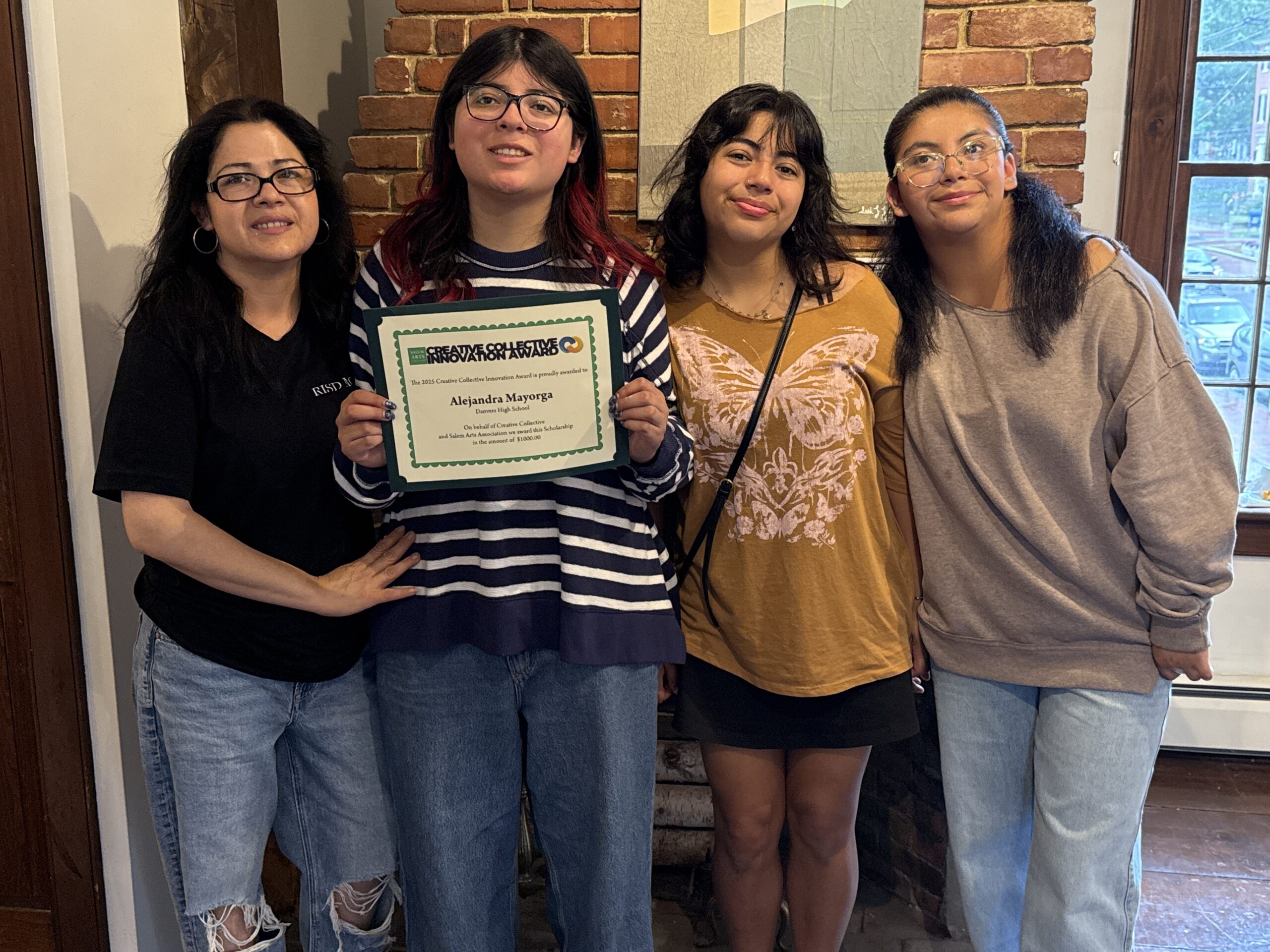 Four smiling community members gather inside as we celebrate Alejandra Mayorga, certificate in hand, against a lively brick backdrop.