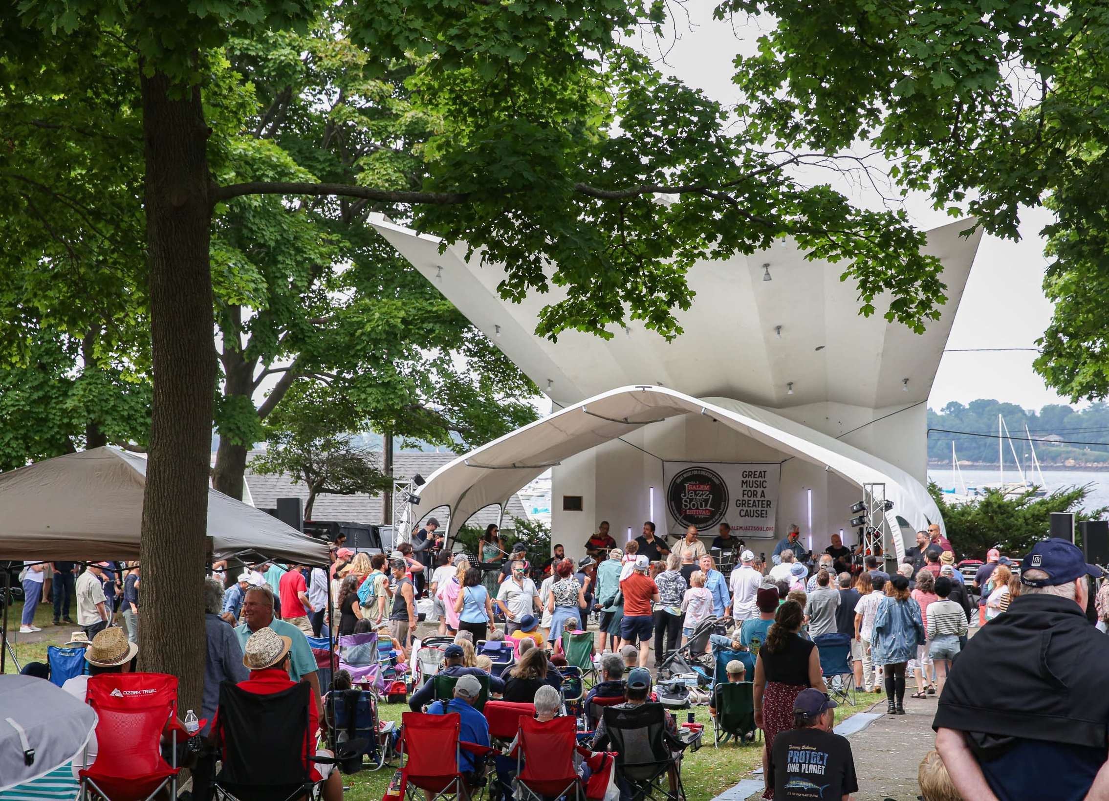 Our vibrant community gathers for a summer concert at Salem Willows—neighbors relax on lawn chairs, kids dance under shady trees, and boats drift by in the sun, all while we enjoy live local music together.