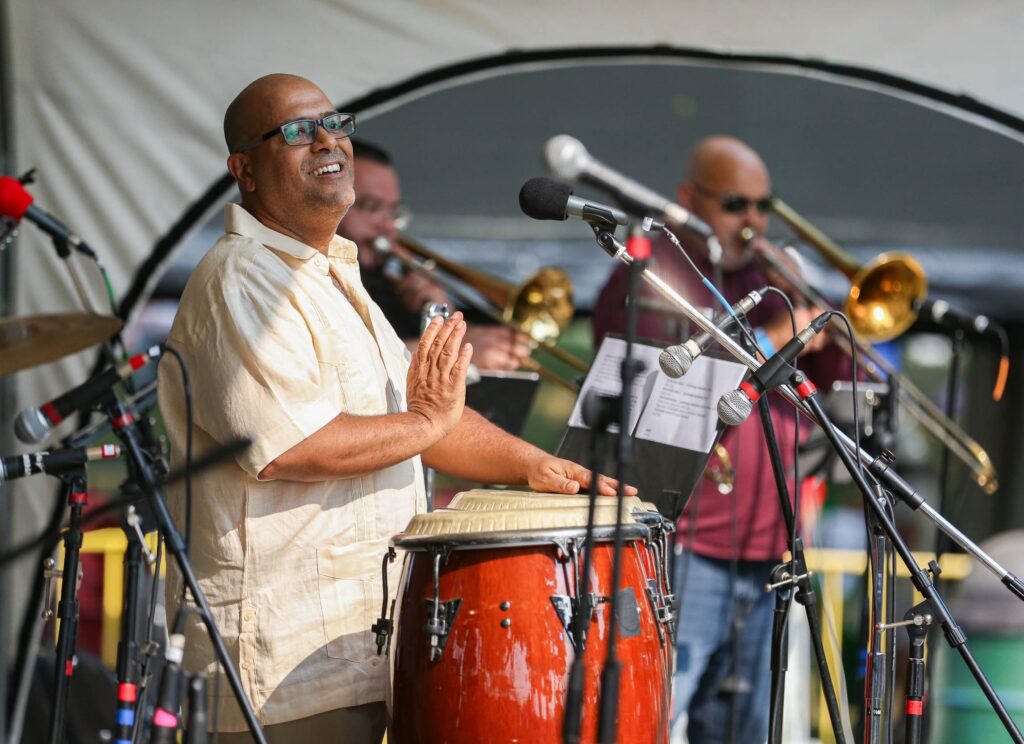 Here’s a revised, SEO-friendly alt text with your requested tone and elements: A joyful local drummer leads our Salem Willows concert, congas pulsing as bandmates on trombones fill the stage with creative energy.