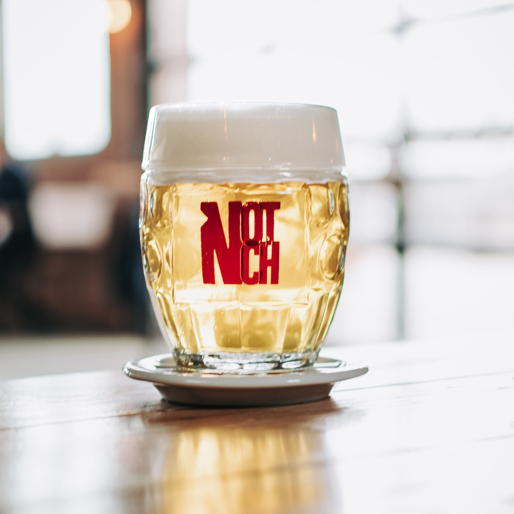 A frosty glass mug of Notch beer with a thick, creamy head and our bold red logo shines on a wooden table—crafted for our community.