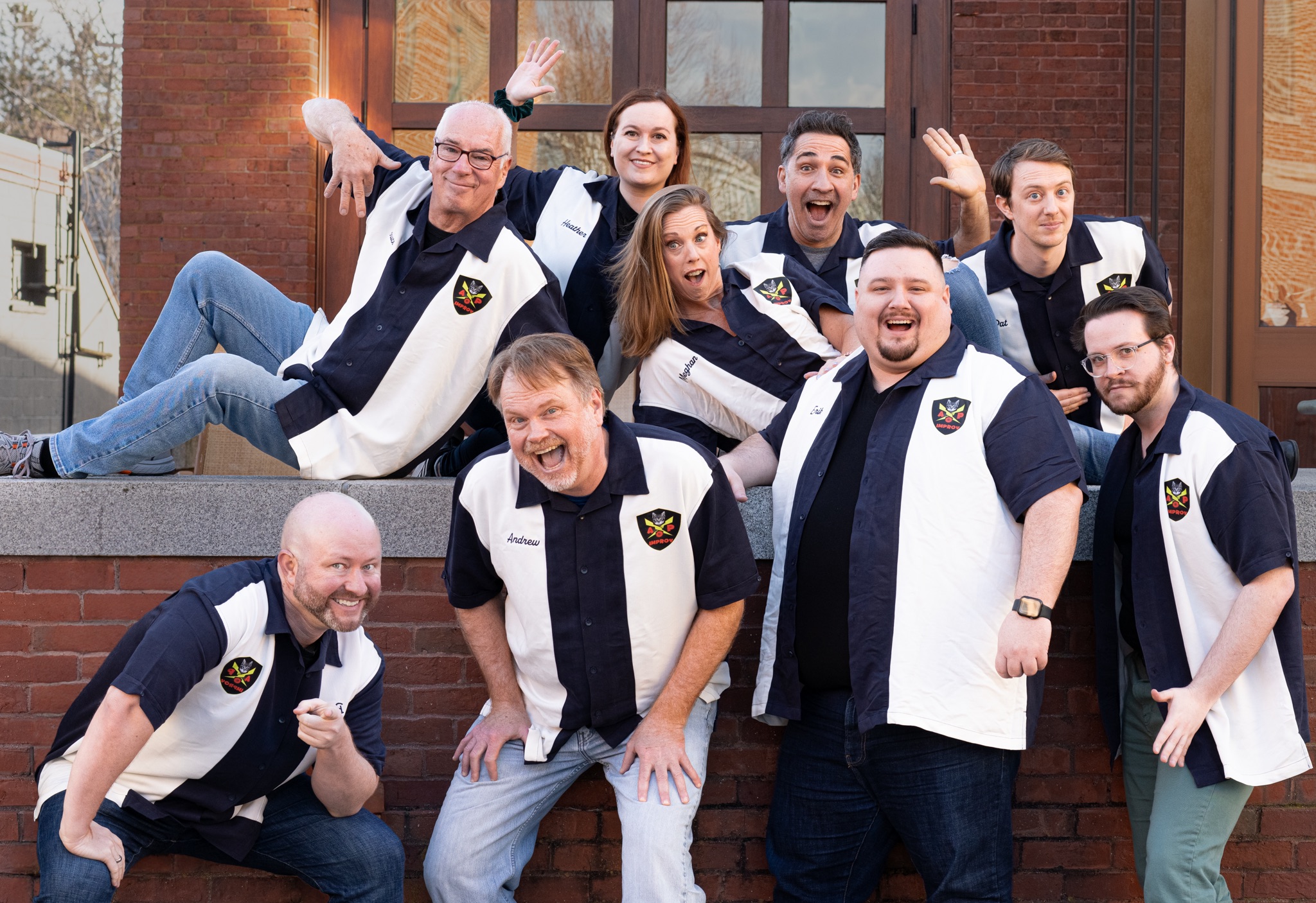 Nine adults in bold black-and-white shirts beam with joy, striking playful poses together outside a brick building—celebrating our creative community spirit.