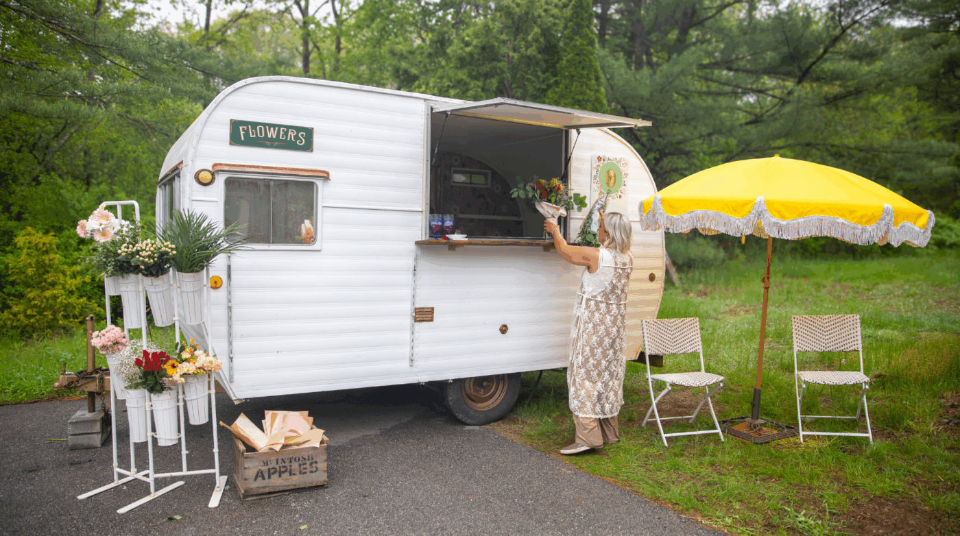 A woman in a lively patterned dress picks fresh blooms at our vintage white flower truck, surrounded by trees, sunshine, and community vibes.
