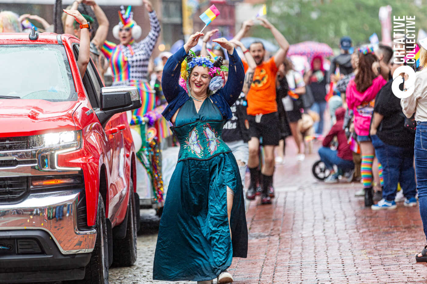 A woman in a teal dress and floral crown twirls with joy on our rain-kissed brick street at Pride, surrounded by bold costumes, rainbow flags, and cheering neighbors—a true celebration of our creative community spirit.
