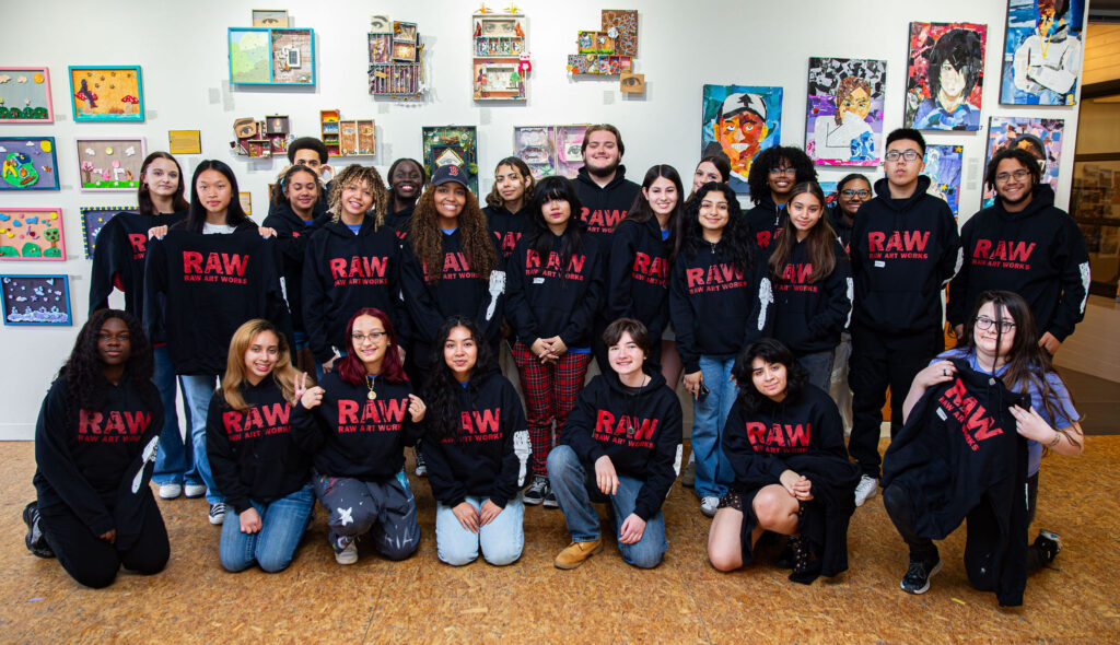 A vibrant crew of young creatives in matching RAW hoodies gathers indoors, beaming before a wall of bold, local art.