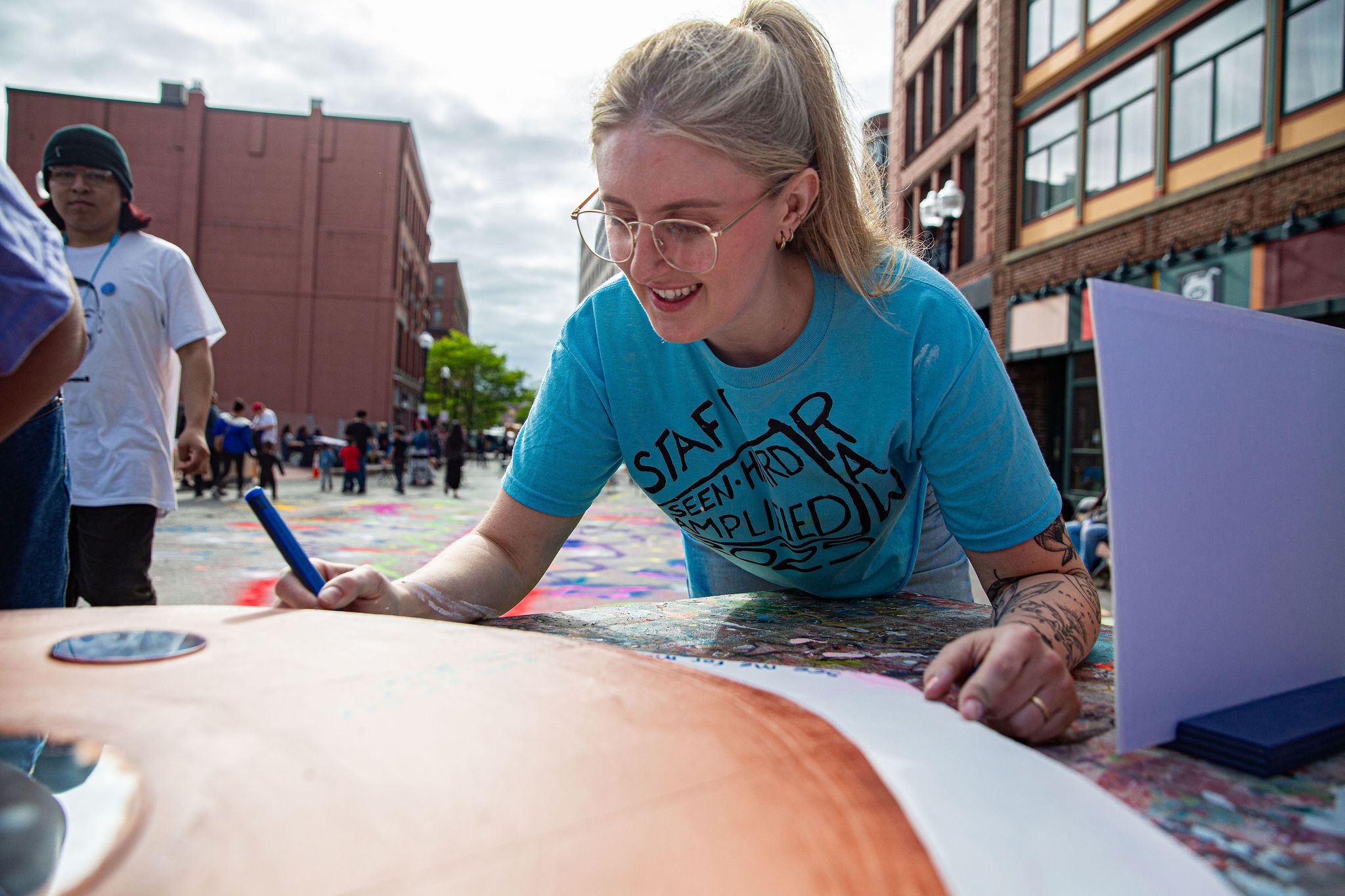 Smiling staffer with glasses and a ponytail brings bold art to life on canvas at our Lynn street art fest, surrounded by colorful community energy.