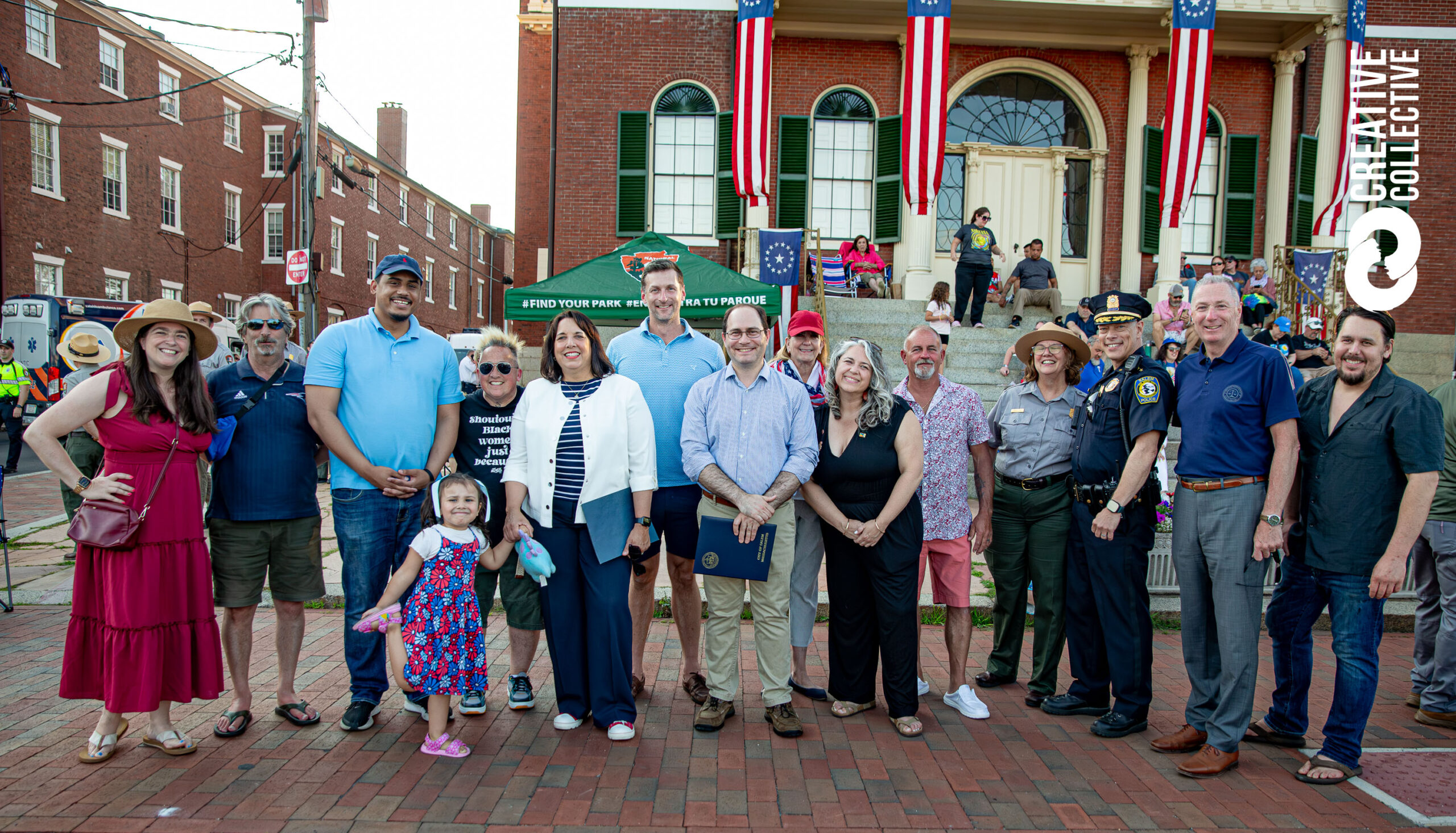 Smiling neighbors of all ages—including officials—gather outside our historic, flag-adorned building, celebrating local pride together. Let me know if the image spotlights artists or events and I can tailor it even further!.
