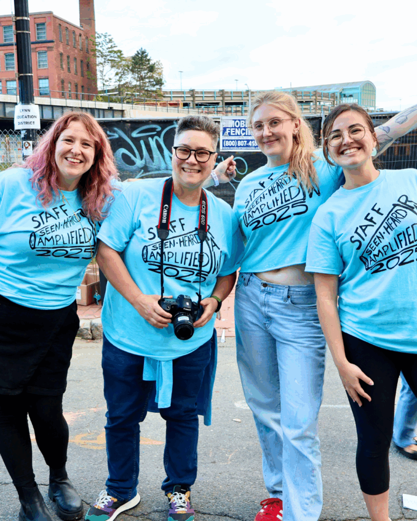 Raw Artworks staff smile together in matching “Staff Seen-Heard Amplified 2025” tees outside a vibrant urban backdrop—camera in hand and creative spirit shining. Lynn MA.