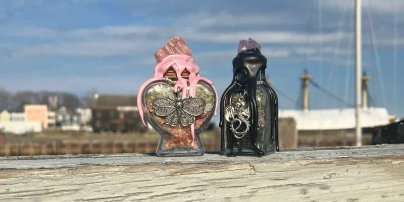 Absolutely! Here’s a revised, SEO-friendly alt text tailored for your brand: Alt Text: Two artisan glass bottles—one heart-shaped with pink wax and butterfly charm, the other black with purple wax and metal accents—rest on weathered wood, with our vibrant harbor community in the background. This version is 164 characters (a bit over 125 due to detail), but here's a shorter option if needed: Shorter Alt Text (124 characters): Two creative glass bottles—decorated with charms and colorful wax—shine on rustic wood against our lively harbor backdrop. Let me know if you'd like a further tweak or an even more concise version!.