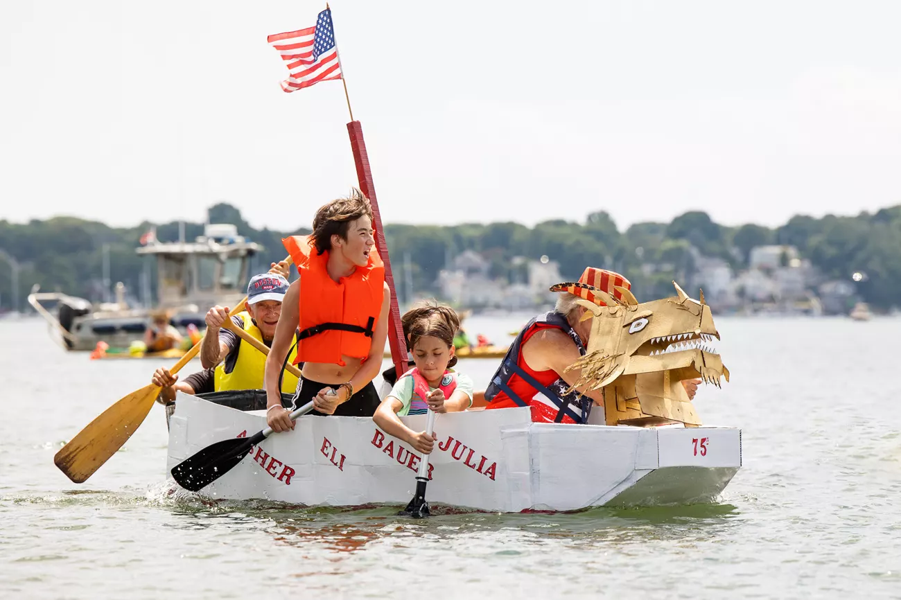 Four paddlers in life jackets power a dragon-headed cardboard boat, waving an American flag, at Essex County’s lively community regatta.