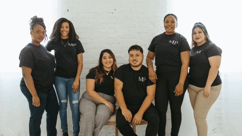 Six creative locals in matching “MP” shirts smile warmly together, radiating Essex County community spirit indoors by a sunlit wall.