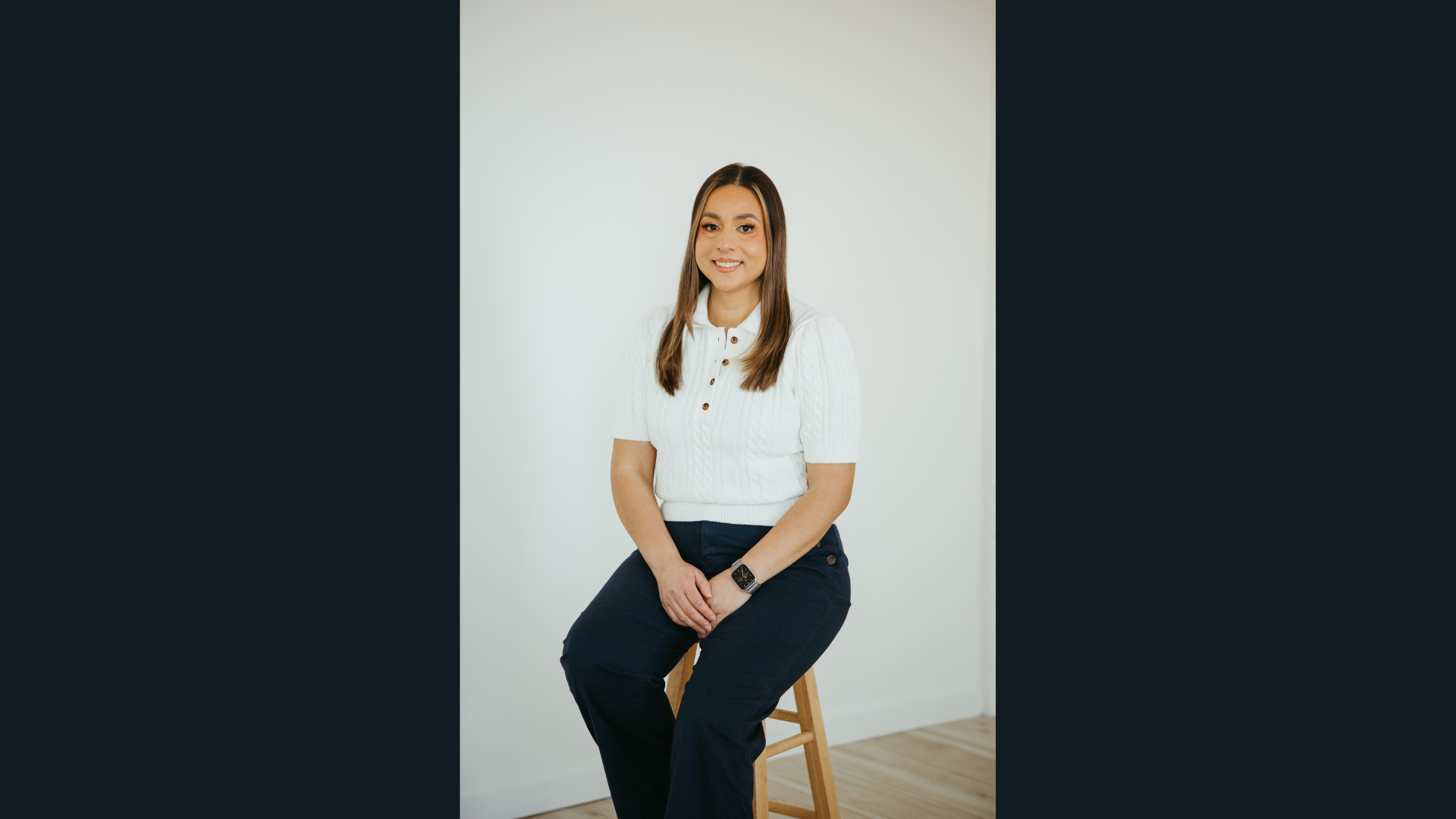 Hope Blooms Floristry’s founder beams creatively, connecting Essex County with blooms, seated on a stool by a white wall.