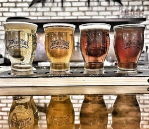 Four vibrant Granite Coast Brewing beers—from pale to amber—shine on a tray, reflecting Essex County’s lively brew scene.