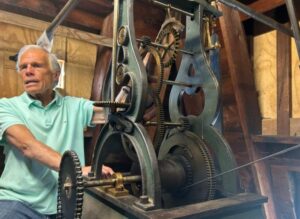 An older man in a light green polo proudly stands by an antique clock’s gears—a symbol of creativity and history in Essex County.