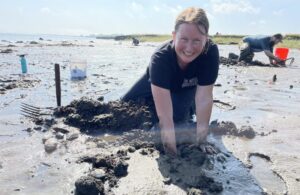 Smiling friends dig in Essex County’s wet sand, tools at hand, building connections as clouds drift above the lively shoreline.