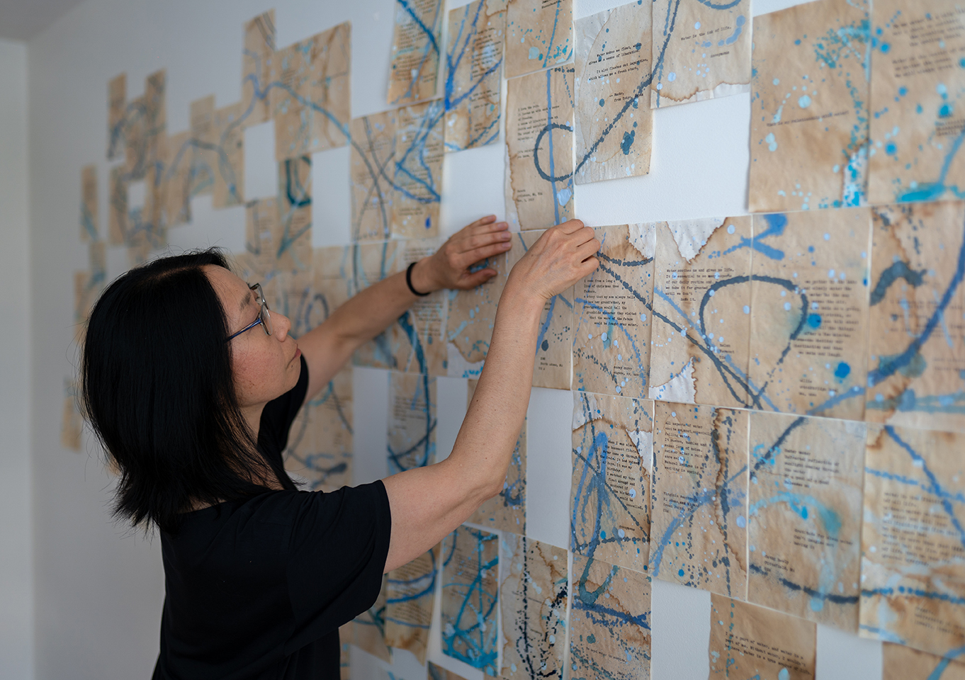 A creative leader in a black shirt arranges vibrant, text-filled papers with blue splatters, inspiring Essex County’s community wall.