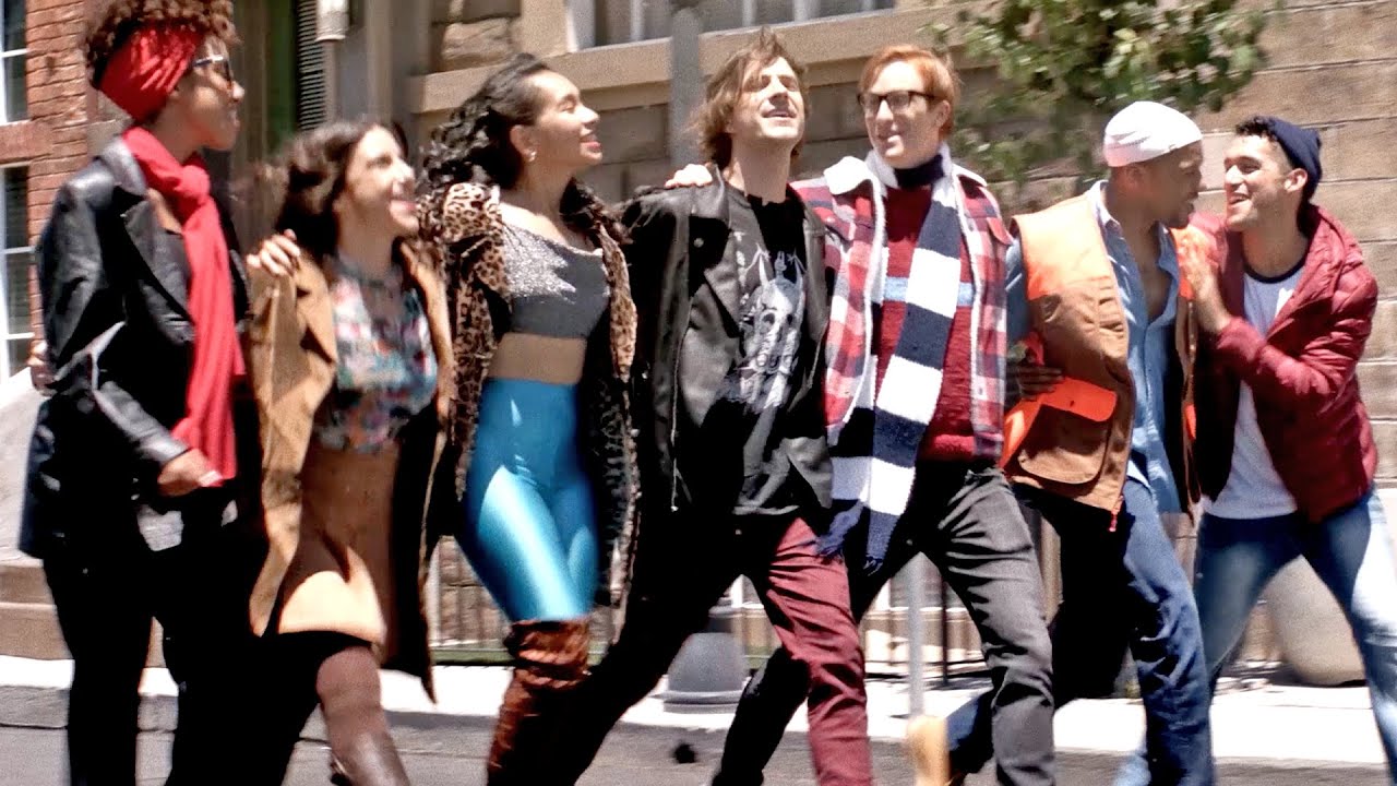 Seven people in vibrant, creative outfits link arms and laugh together outdoors, radiating Essex County’s warm community spirit.