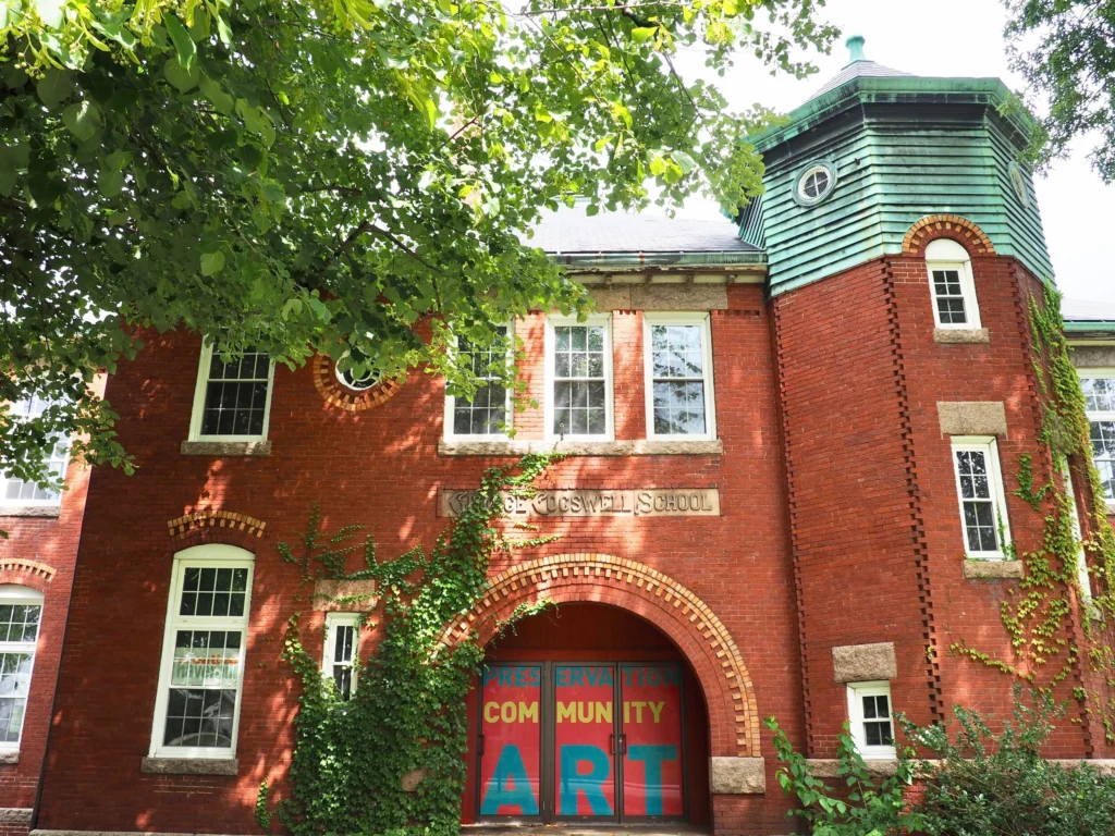 A lively red brick building with arched windows and a green tower bursts with “Preservation Community Art”—Essex County’s creative hub, wrapped in vibrant ivy and shaded by leafy trees. Creative Haverhill