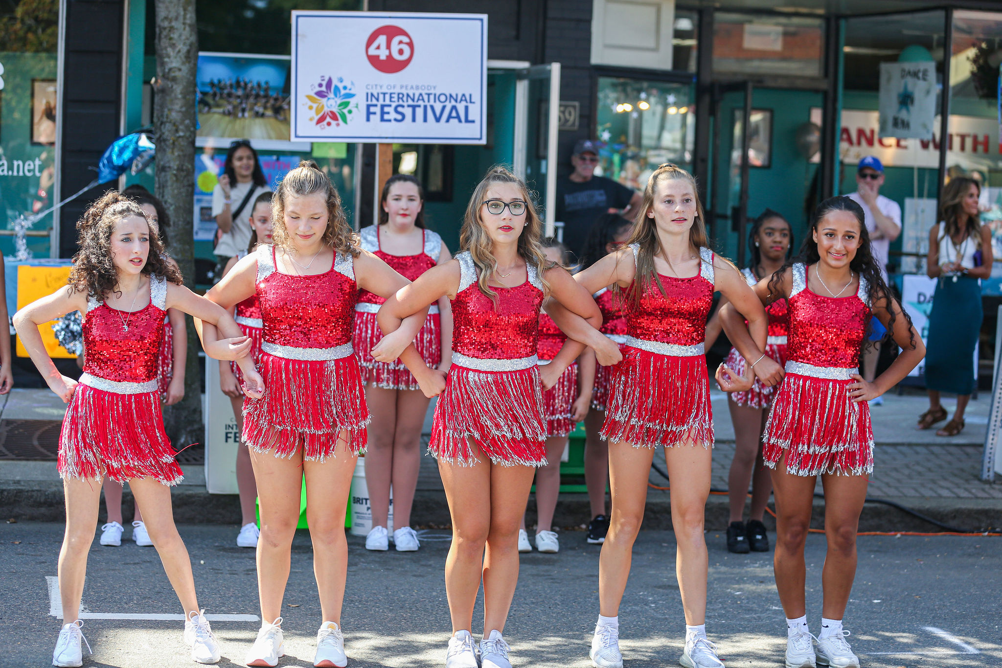 Five girls shine in red sequins, striking poses together on stage at Peabody International Festival, uniting Essex County in joy.