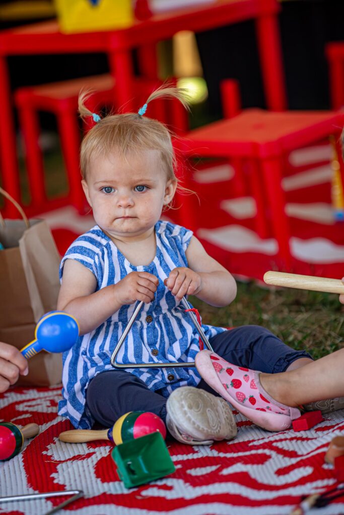 In a lively play space, a joyful toddler makes music on a triangle, surrounded by vibrant toys and bold red décor, sparking Essex County creativity.