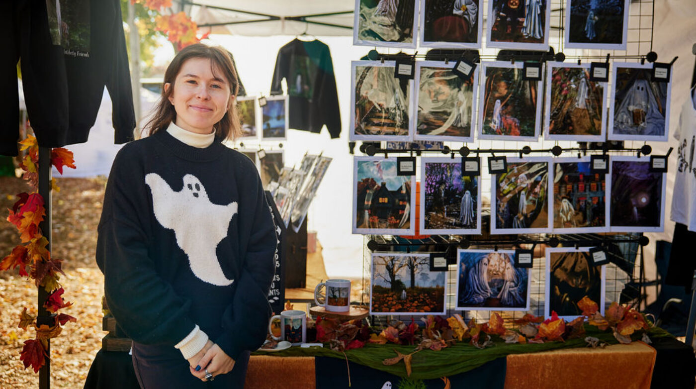 A creative local vendor beams in front of an autumn-themed Essex County market booth, vibrant with Halloween art and community spirit.