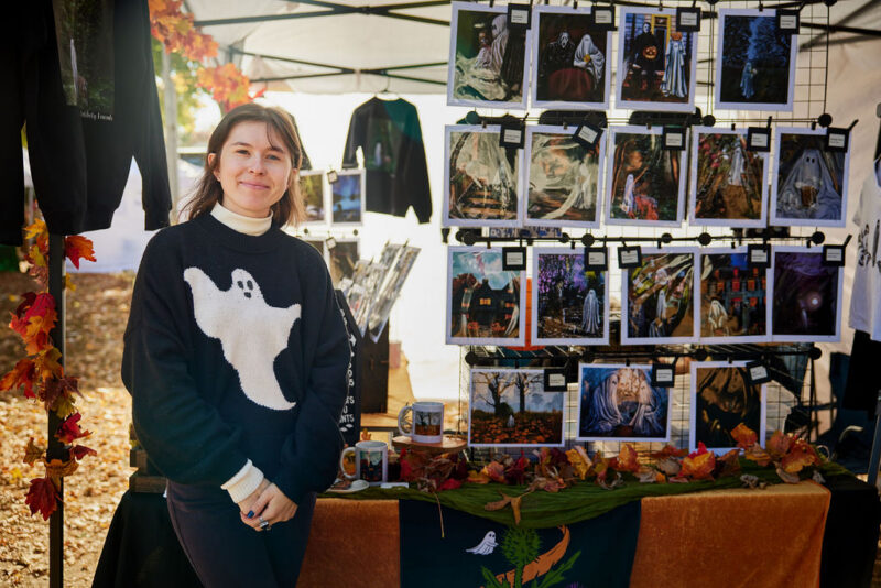 A creative local vendor beams in front of an autumn-themed Essex County market booth, vibrant with Halloween art and community spirit.
