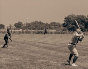 A sepia photo captures community spirit as a pitcher fires to a batter, with Essex County fields and crops framing the lively game.