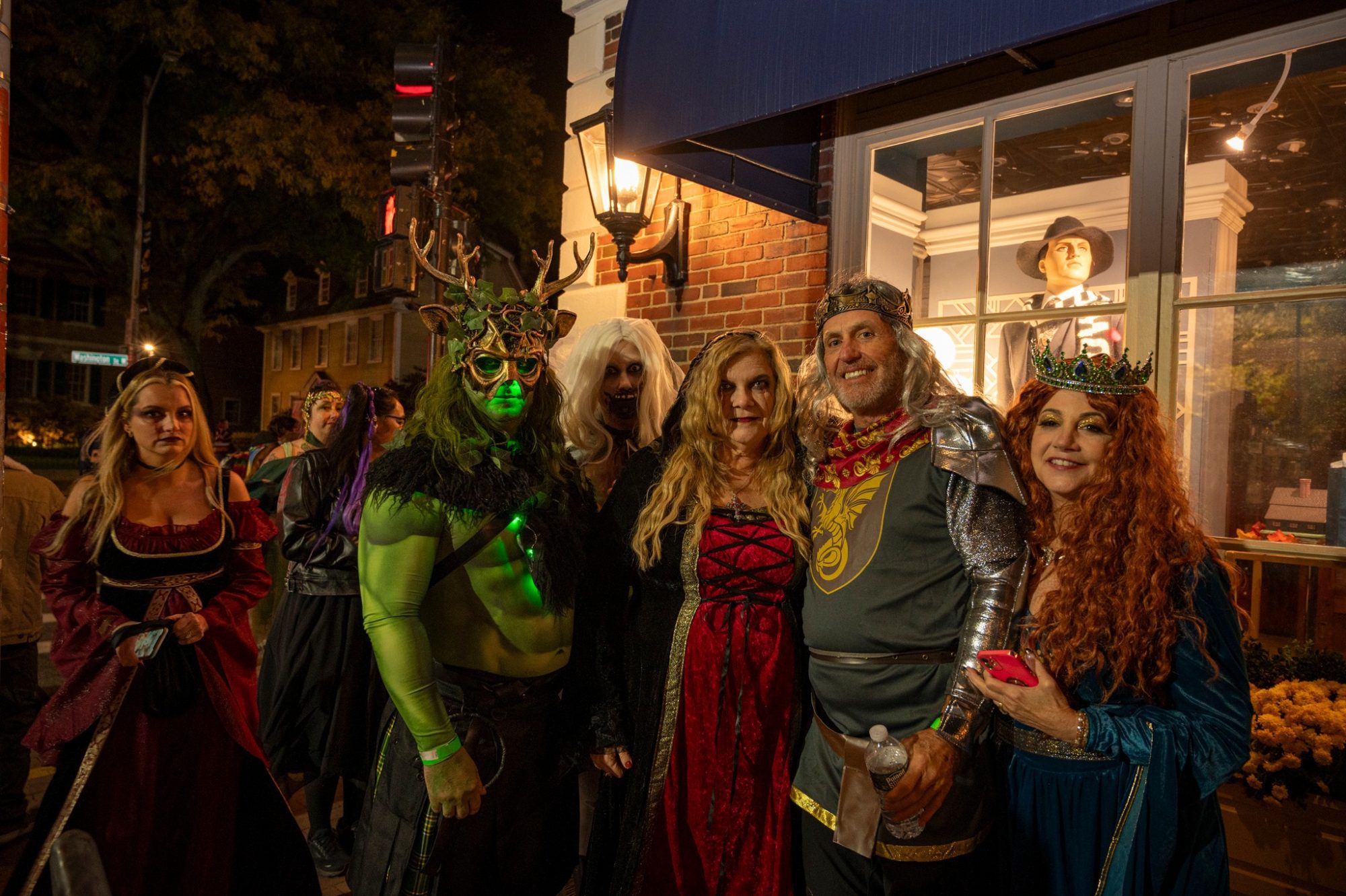 Six adults in vibrant medieval and fantasy costumes gather joyfully outside a lively Essex County shop at night, celebrating local creativity.