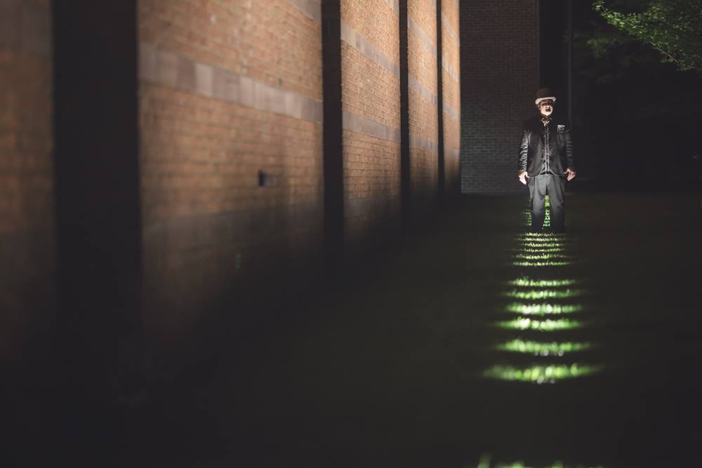 In Essex County, a creative soul in a hat stands by a brick wall at night, as vibrant lights connect the community in glowing unity.