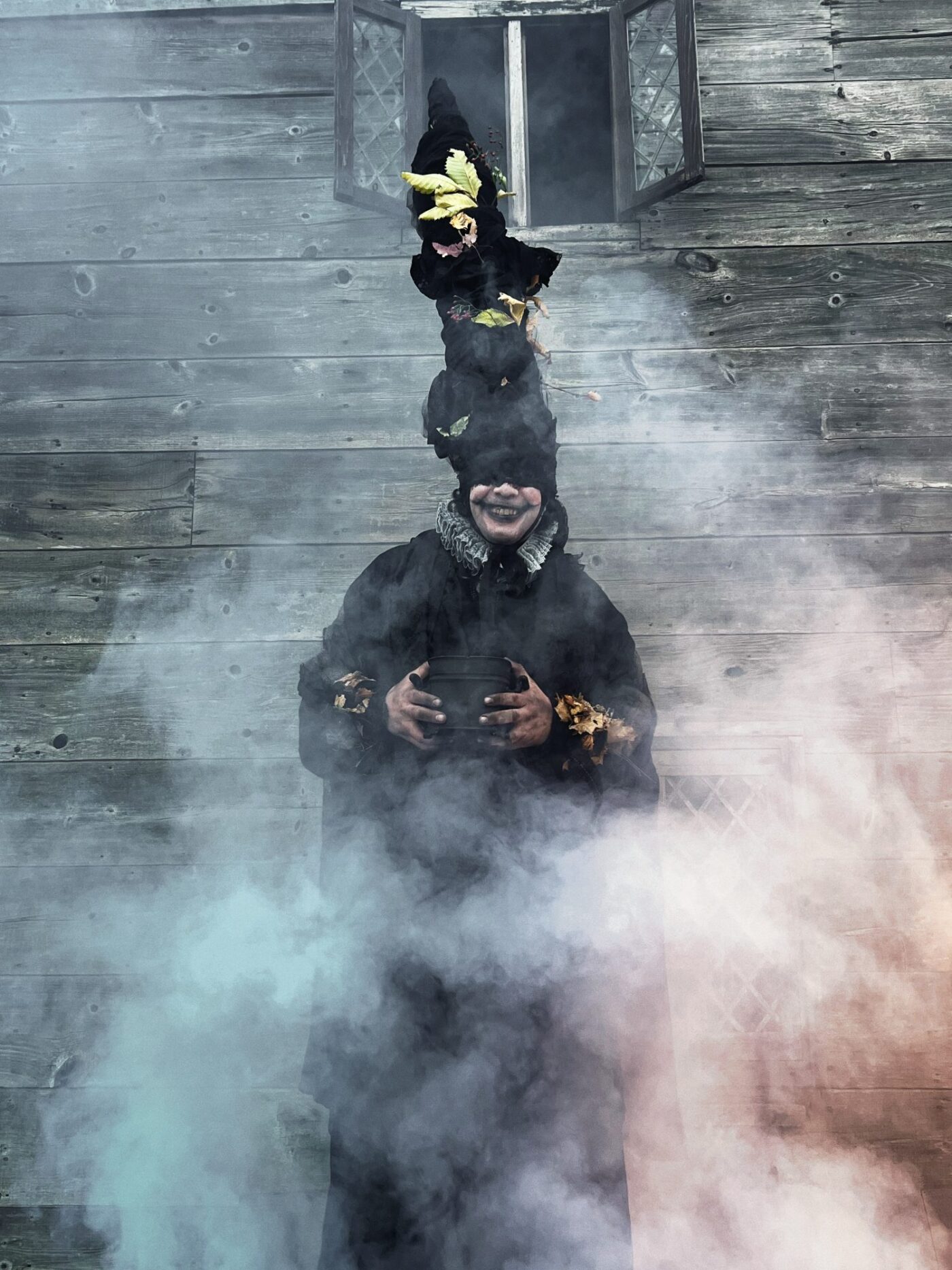 A creative figure in a worn black robe and tall hat brings cauldron magic to a lively local hub, mist swirling as neighbors gather nearby.